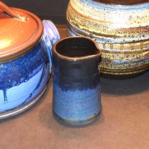 May include: A small ceramic pitcher with a dark black upper section and a textured blue lower base, positioned between a large blue lidded ceramic pot and a round, multi-colored ceramic vessel with horizontal brown, white, and blue bands.