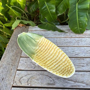 May include: A ceramic corn-shaped serving dish. The dish is yellow with a textured surface, mimicking corn kernels, and has a green husk-like end. It rests on a weathered wooden surface, with green foliage in the background.