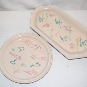 May include: Two ceramic serving dishes. One is round, the other is rectangular with angled sides. Both are off-white with colorful abstract designs in blue, pink, and green. The dishes are likely for serving food.