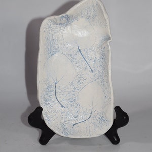 May include: A white ceramic tray with a textured surface and a leaf design in light blue. The tray is rectangular with an irregular edge and is displayed on a black stand. Decorative home decor.