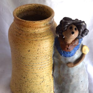 May include: A ceramic vessel featuring a speckled yellow cylindrical vase attached to a sculpted figure. The figure has dark brown hair, a blue collar, and a light gray garment. The vase has a wide opening and a textured, speckled surface finish.