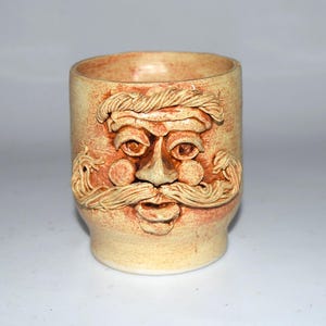 May include: A cylindrical ceramic mug with a light tan, speckled finish. The exterior features a three-dimensional sculpted face with a prominent mustache, bushy eyebrows, round cheeks, and a small mouth, all integrated into the clay surface.