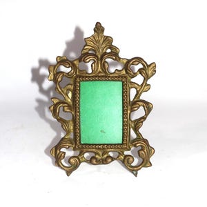 May include: A rectangular picture frame made of antique brass with an ornate, scrolling leaf-like border and a beaded inner edge. The center of the frame contains a solid, bright green rectangular insert, creating a high-contrast visual effect.