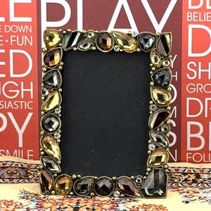 May include: A rectangular picture frame featuring a dark metal border adorned with an array of faceted, multi-shaped amber, brown, and clear glass crystals. The center of the frame is a solid black rectangular insert for displaying a photograph.