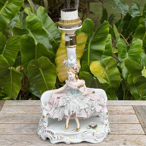 May include: A vintage porcelain lamp base featuring a seated figurine in a pink and blue dress. The lamp has a white and gold finish, with a decorative base and a light socket at the top. The figurine is holding a fan.