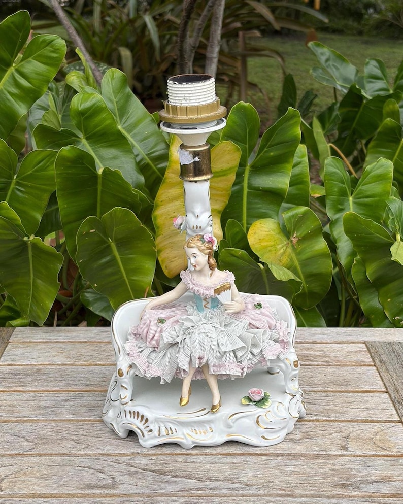 May include: A vintage porcelain lamp base featuring a seated figurine in a pink and blue dress. The lamp has a white and gold finish, with a decorative base and a light socket at the top. The figurine is holding a fan.