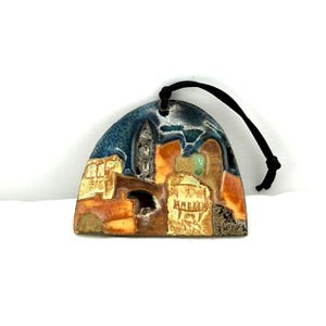 May include: A ceramic ornament depicting a cityscape with a blue sky, orange buildings, and a black cord for hanging. The ornament has a textured surface and a unique, artistic design.
