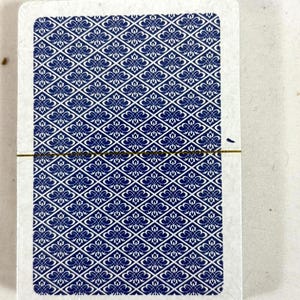 Puede incluir: Una baraja de cartas con un patr&oacute;n floral azul y blanco en el reverso. Las cartas est&aacute;n selladas en pl&aacute;stico transparente, con una banda dorada. Los bordes de las cartas son blancos.