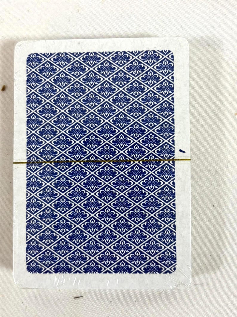 Puede incluir: Una baraja de cartas con un patr&oacute;n floral azul y blanco en el reverso. Las cartas est&aacute;n selladas en pl&aacute;stico transparente, con una banda dorada. Los bordes de las cartas son blancos.