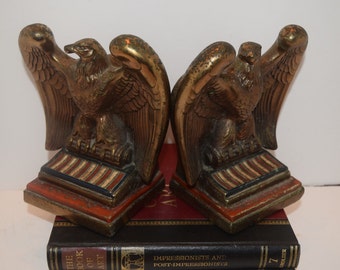 May include: A pair of gold eagle bookends with red, white, and blue striped bases. The bookends are sitting on top of a stack of books.