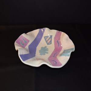 May include: A white ceramic bowl with a wavy rim. The bowl is decorated with a geometric pattern in shades of purple, pink, and blue.