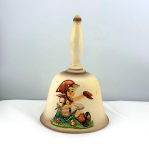 May include: A vintage ceramic bell with a cream-colored finish and a wooden handle. The bell features a painted illustration of a girl in a red headscarf and blue dress, sitting in a field of flowers. The bell is approximately 6 inches tall.