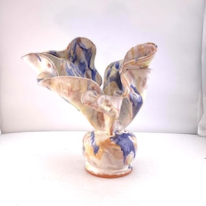 May include: A handcrafted ceramic vase with a unique, abstract design. The vase features a bulbous base and flared, ruffled top. The color palette includes shades of blue, peach, and brown, creating a marbled effect. The vase is a decorative art piece.