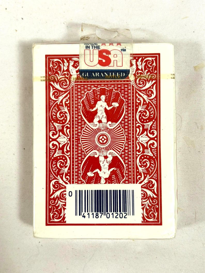 Puede incluir: Una baraja de cartas sellada con un dise&ntilde;o rojo y blanco. El reverso de la carta presenta un patr&oacute;n sim&eacute;trico con una figura central y detalles ornamentados. El embalaje incluye el texto "MADE IN THE USA" y un c&oacute;digo de barras.