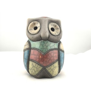 May include: A ceramic owl figurine with a gray head and body, featuring large, round eyes. The owl's chest is decorated with colorful, geometric shapes in blue, pink, green, and yellow. The figurine has a crackle glaze finish.