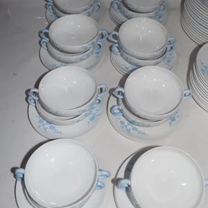 Pu&ograve; includere: Una collezione di zuppiere bianche con manici azzurri e piattini abbinati. I piattini e le ciotole sono decorati con un delicato motivo floreale blu. Una pila di piatti abbinati &egrave; visibile sullo sfondo.