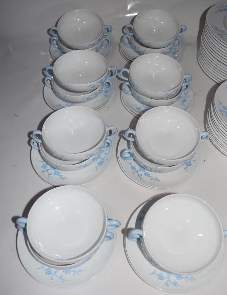 Pu&ograve; includere: Una collezione di zuppiere bianche con manici azzurri e piattini abbinati. I piattini e le ciotole sono decorati con un delicato motivo floreale blu. Una pila di piatti abbinati &egrave; visibile sullo sfondo.