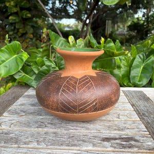 May include: A round terracotta vase with a wide, flared rim and a smooth, light brown interior. The exterior features a dark, mottled brown finish with a series of etched, leaf-shaped patterns radiating from the neck down to the base.