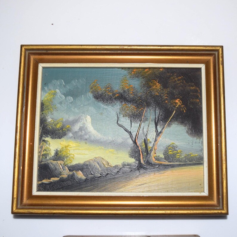 Op de afbeelding: Olieverfschilderij van een landschap in een gouden lijst. Het kunstwerk toont een boom met donkergroene en bruine bladeren, een berg en een bewolkte lucht. Het schilderij heeft een textuur.
