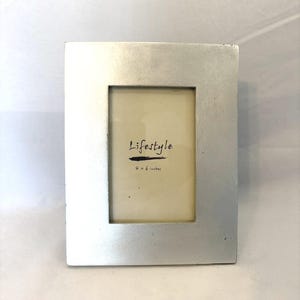 May include: A rectangular silver picture frame with a cream-colored interior. The frame has the word "Lifestyle" and the dimensions "4 x 6 inches" printed on the inside. The frame is made of a metallic material.