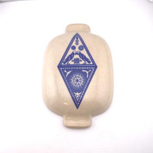 May include: Cream-colored ceramic flask with a blue diamond-shaped design. The design features two birds, floral elements, and a sunburst pattern. The flask has a rounded body and a small, cylindrical neck.