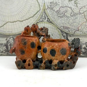 May include: A pair of carved stone containers with a reddish-orange hue and black spots. The containers are connected by a dark brown, intricately carved base, set against a vintage map background. The containers are likely for decorative purposes.