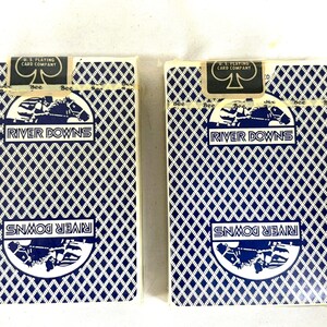 Puede incluir: Dos barajas de cartas con un patr&oacute;n de diamantes azules y las palabras "River Downs" en una pancarta blanca. La parte superior de las barajas de cartas tiene un logotipo en blanco y negro que dice "U.S. Playing Card Company".
