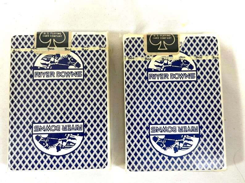 Puede incluir: Dos barajas de cartas con un patr&oacute;n de diamantes azules y las palabras "River Downs" en una pancarta blanca. La parte superior de las barajas de cartas tiene un logotipo en blanco y negro que dice "U.S. Playing Card Company".