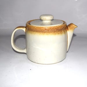 May include: A ceramic teapot with a cream-colored body and a speckled finish. The teapot features a brown glazed rim around the top edge and a matching lid with a small knob handle. It has a curved handle on the left and a short spout on the right.