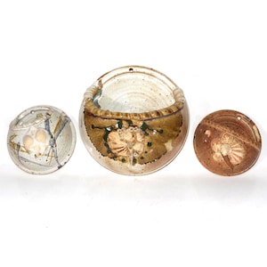 May include: Three ceramic pieces: a small round bowl, a larger bowl with a handle, and a flat, round disc. The pieces are earth-toned with floral designs in brown, beige, and green. Each piece has a hole for hanging.