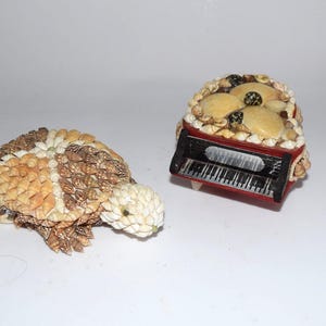 May include: Two decorative items: a turtle figurine and a miniature piano. The turtle is made of small, tan and white shells. The piano is red with a black keyboard and is decorated with shells and cream-colored accents.