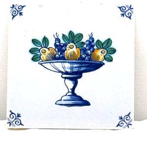 May include: Square ceramic tile with a hand-painted design. The tile features a blue and white color scheme, with a central image of a fruit bowl filled with yellow fruit and blue grapes. Decorative blue corner accents complete the design.