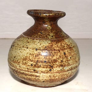 May include: A small, round ceramic vase with a narrow neck and a flared rim. The surface features a speckled, mottled glaze in shades of tan, beige, and dark brown, with horizontal incised lines circling the lower body of the vessel.