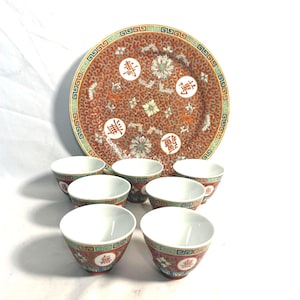 May include: A vintage Chinese porcelain tea set featuring a large plate and six small teacups. The plate and cups have a red background with floral and Chinese character designs. The rims are decorated with a green and gold pattern.
