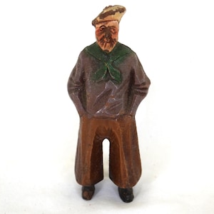 Könnte beinhalten: Kleine, handgeschnitzte Holzfigur eines Seemanns. Der Seemann trägt ein braunes Hemd, eine braune Hose, einen grünen Schal und einen beigen Hut. Die Figur hat ein detailliertes Gesicht mit Bart und Schnurrbart. Die Hände des Seemanns sind in seinen Taschen.