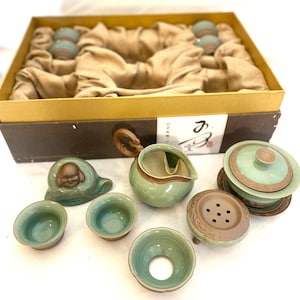 May include: A complete tea set with a light green and brown color scheme. The set includes a teapot, cups, and a tea strainer. The tea set is presented in a brown box with a gold-colored trim. The set includes a small ceramic Buddha statue.
