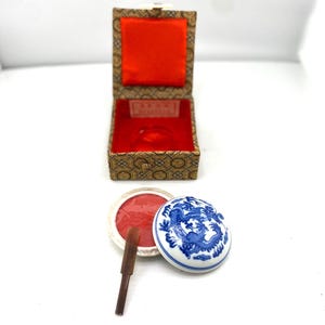 May include: An open, ornate box with a red interior and a small, round, red ink pad. A small, brown applicator and a white and blue ceramic ink pot with a dragon design are also present.