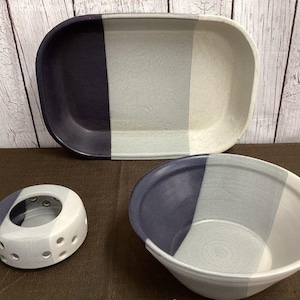 May include: A set of three ceramic pieces: a rectangular platter, a bowl, and a small, perforated candle holder. Each piece is divided into three vertical sections of navy blue, light gray, and white. The items are arranged on a brown surface.