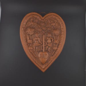 May include: A heart-shaped, brown wooden wall hanging with carved relief details. The relief depicts figures, birds, and foliage. The bottom of the heart has a smaller heart with the letters "RIO" carved into it. A small loop is at the top for hanging.