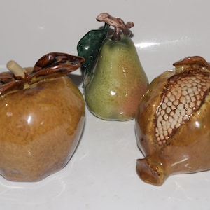 May include: Three ceramic fruit sculptures, an apple, a pear, and a pomegranate, in shades of brown and green.