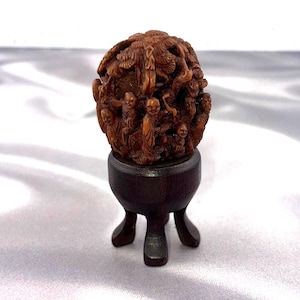 May include: A brown, intricately carved wooden netsuke featuring multiple figures and foliage, mounted on a dark brown, three-legged wooden stand. The carving displays detailed human faces and organic textures throughout the spherical shape.