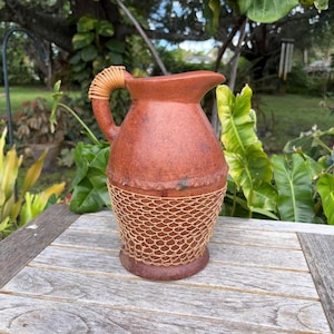 May include: A terracotta-colored pitcher with a woven handle and a woven net design around the base. The pitcher has a spout for pouring and sits on a wooden surface. The pitcher is a decorative item.