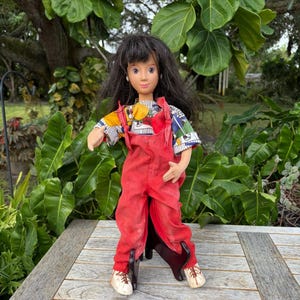 May include: A vintage doll with long black hair, wearing a colorful patterned shirt and red overalls. The doll has a fair complexion and is wearing white shoes. The doll is standing on a wooden surface with a green leafy background.