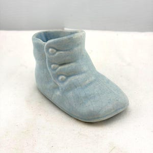 May include: A light blue ceramic baby bootie with a crackle glaze finish. The boot has three button details on the side and a rounded toe. The boot is a decorative item.