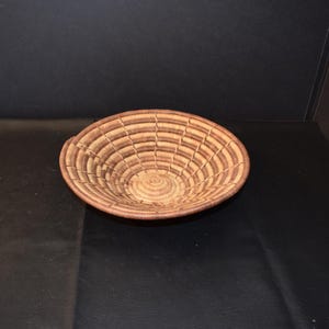 May include: A round, woven basket with a shallow bowl shape. The basket features concentric circles of light and dark brown fibers, creating a textured pattern. The rim is a darker shade of brown. The basket is likely handmade and suitable for decorative or functional use.