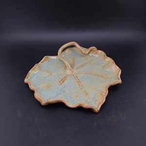 May include: A ceramic leaf-shaped dish with a light blue glaze and brown accents. The dish has a handle and a textured surface, with a brown rim. The leaf design is detailed with vein-like patterns.