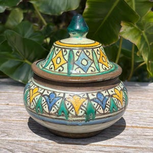 May include: A small, ceramic jar with a lid. The jar is decorated with geometric patterns in yellow, blue, and green against a cream background. The lid has a green finial. The jar sits on a wooden surface.