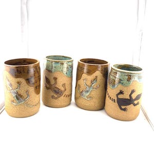 May include: Four ceramic tumblers with a tan base and brown rim. Each tumbler features a lizard design in various colors, including blue, brown, and black. The top of each tumbler has a turquoise glaze.
