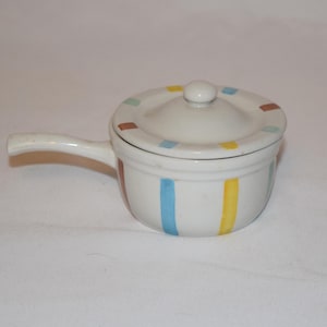 May include: A small, white ceramic pot with a lid and handle. The pot is decorated with vertical blue and yellow stripes, and the lid has a ring of small, colorful squares. A classic kitchenware item.