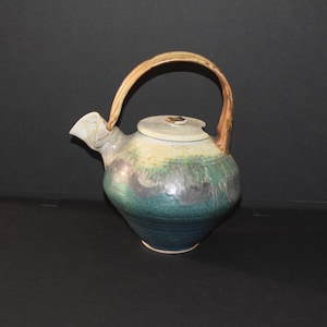 May include: A handmade ceramic teapot with a brown handle and a green and blue glaze. The teapot has a round body and a lid with a small knob.